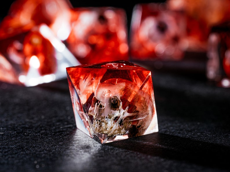 Puede incluir: Un dado poli&eacute;drico transl&uacute;cido de m&uacute;ltiples facetas hecho de resina roja. Dentro del material transparente de color rojo intenso, hay incrustado un cr&aacute;neo humano en miniatura detallado y realista. El dado tiene bordes geom&eacute;tricos.