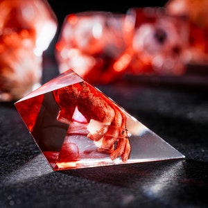 Puede incluir: Un dado de resina triangular y transl&uacute;cido que presenta una inclusi&oacute;n de color rojo intenso de aspecto org&aacute;nico suspendida en el material transparente. El objeto de bordes afilados y m&uacute;ltiples facetas capta la luz, revelando un n&uacute;cleo complejo.