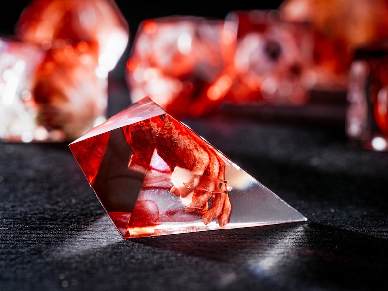Puede incluir: Un dado de resina triangular y transl&uacute;cido que presenta una inclusi&oacute;n de color rojo intenso de aspecto org&aacute;nico suspendida en el material transparente. El objeto de bordes afilados y m&uacute;ltiples facetas capta la luz, revelando un n&uacute;cleo complejo.