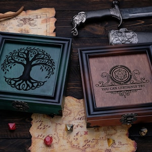 May include: Two square wooden boxes with ornate frames and metal latches. One box is green with a black tree silhouette, the other is brown with a black engraved 20-sided die icon and the text "YOU CAN CERTAINLY TRY" surrounded by decorative scrollwork.
