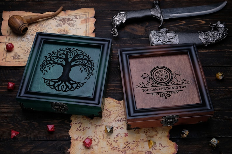 May include: Two square wooden boxes with ornate frames and metal latches. One box is green with a black tree silhouette, the other is brown with a black engraved 20-sided die icon and the text "YOU CAN CERTAINLY TRY" surrounded by decorative scrollwork.