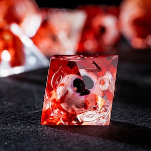 Puede incluir: Un dado poli&eacute;drico rojo transl&uacute;cido con un globo ocular humano realista incrustado en la resina. El dado presenta bordes afilados y n&uacute;meros grabados claramente en sus caras, con otros dados similares desenfocados en el fondo.