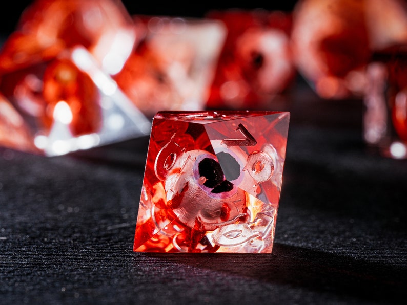 Puede incluir: Un dado poli&eacute;drico rojo transl&uacute;cido con un globo ocular humano realista incrustado en la resina. El dado presenta bordes afilados y n&uacute;meros grabados claramente en sus caras, con otros dados similares desenfocados en el fondo.