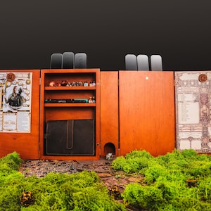 Könnte beinhalten: Ein hölzerner Tisch-Gaming-Organizer mit mehreren Fächern. Der Organizer ist geöffnet und zeigt Regale mit Würfeln, einem Stift und einem Notizbuch. Die linke Seite zeigt ein Charakterblatt und Kunstwerke. Der Hintergrund ist dunkel.