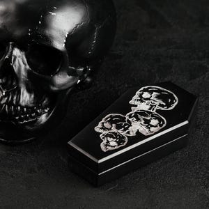 Puede incluir: Una caja negra con forma de ata&uacute;d que presenta un gr&aacute;fico plateado de cuatro calaveras apiladas en la tapa, colocada junto a una gran escultura de calavera negra lisa. La caja tiene una base rectangular y una tapa con bordes definidos.