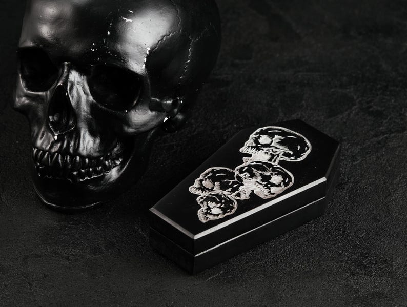 Puede incluir: Una caja negra con forma de ata&uacute;d que presenta un gr&aacute;fico plateado de cuatro calaveras apiladas en la tapa, colocada junto a una gran escultura de calavera negra lisa. La caja tiene una base rectangular y una tapa con bordes definidos.