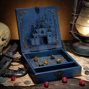 May include: A blue wooden dice box with a hinged lid featuring an engraved castle illustration and metal corner accents. The interior is lined with grey felt and contains several multi-sided dice. A metal chain holds the lid open.