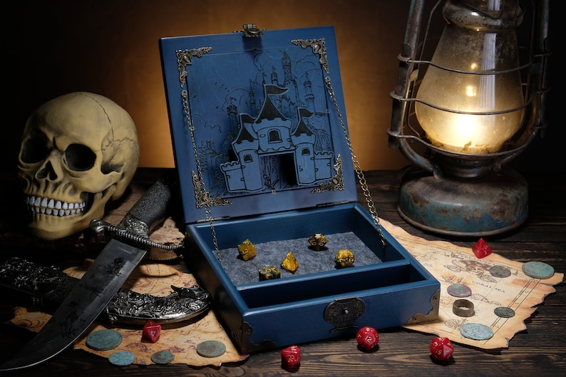 May include: A blue wooden dice box with a hinged lid featuring an engraved castle illustration and metal corner accents. The interior is lined with grey felt and contains several multi-sided dice. A metal chain holds the lid open.