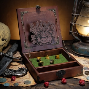 May include: A dark brown wooden dice box with a hinged lid featuring an engraved multi-eyed monster. The interior base is lined with green felt, containing five multi-coloured polyhedral dice. The box has ornate metal corner accents and a latch.