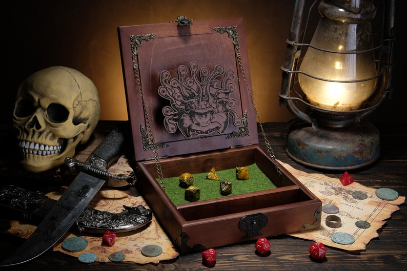 May include: A dark brown wooden dice box with a hinged lid featuring an engraved multi-eyed monster. The interior base is lined with green felt, containing five multi-coloured polyhedral dice. The box has ornate metal corner accents and a latch.