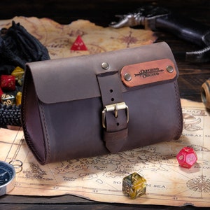 May include: A dark brown leather pouch with a flap closure secured by a brass-toned metal buckle. A rectangular leather patch is attached to the front flap, featuring the embossed text Dungeons & Dragons with a sword graphic and two brass rivets.