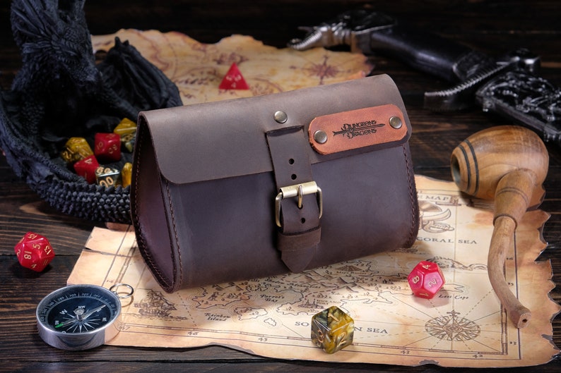May include: A dark brown leather pouch with a flap closure secured by a brass-toned metal buckle. A rectangular leather patch is attached to the front flap, featuring the embossed text Dungeons & Dragons with a sword graphic and two brass rivets.