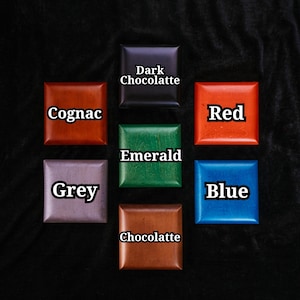 May include: Seven square wooden blocks arranged in a grid, each labelled with a colour name: Dark Chocolatte, Cognac, Red, Emerald, Grey, Blue, and Chocolatte. The blocks feature smooth, polished surfaces in distinct shades of brown, red, green, grey, and blue.