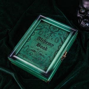 May include: A green wooden box with the words "Alchemy Book" on the lid. The box is decorated with etched illustrations of crystals, bottles, and other mystical symbols. The box has a dark green frame and a brass clasp.