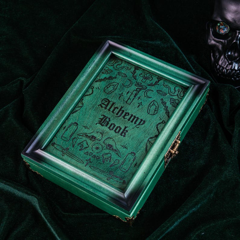 May include: A green wooden box with the words "Alchemy Book" on the lid. The box is decorated with etched illustrations of crystals, bottles, and other mystical symbols. The box has a dark green frame and a brass clasp.