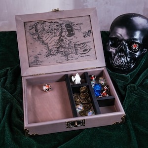 May include: A wooden gaming box with a detailed map of Middle Earth on the lid. Inside, compartments hold dice, miniature figurines, and gold-colored coins. A black skull with a die in its eye rests nearby.
