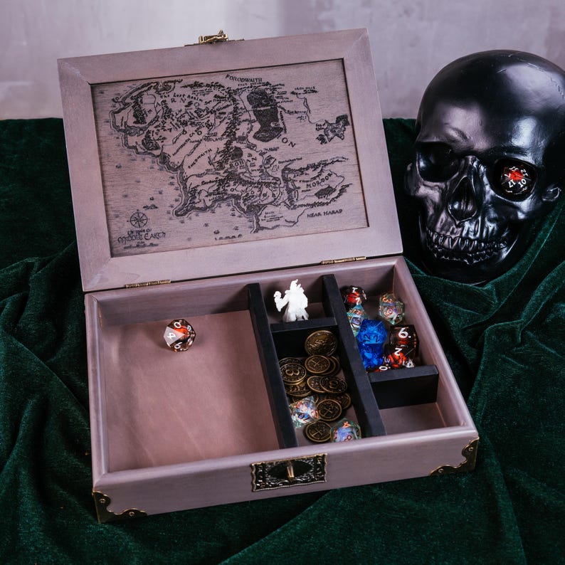 May include: A wooden gaming box with a detailed map of Middle Earth on the lid. Inside, compartments hold dice, miniature figurines, and gold-colored coins. A black skull with a die in its eye rests nearby.
