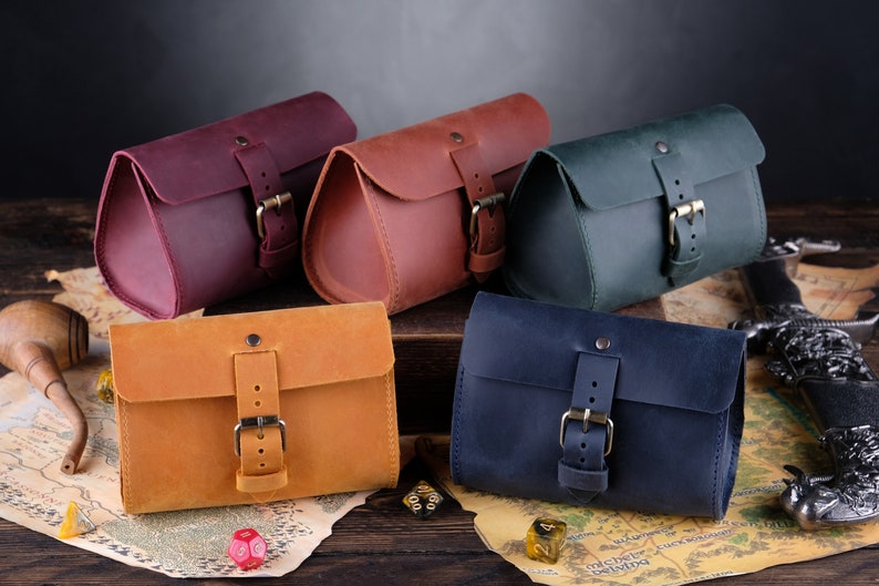 May include: Five rectangular leather pouches with rounded sides, each featuring a single flap secured by a metal buckle strap. The pouches are colored in shades of deep purple, reddish-brown, dark green, mustard yellow, and navy blue.