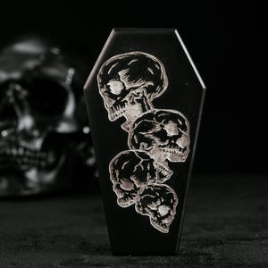 May include: Black coffin-shaped decorative item with four etched skulls. The skulls are stacked vertically, with detailed shading. A blurred black skull is in the background, creating a gothic theme.