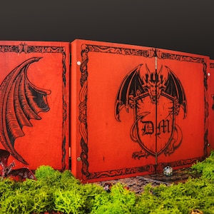 May include: A three-panel red wooden screen featuring black engraved dragon wing designs on the side panels and a central dragon emblem holding a shield with the letters DM. The screen is surrounded by green moss and small game dice.