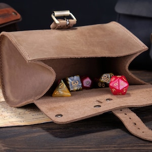 May include: A light brown leather pouch with a buckle closure containing several multi-sided gaming dice. The pouch is open, revealing a red twenty-sided die in the foreground and other assorted dice inside, including yellow and marbled patterns.