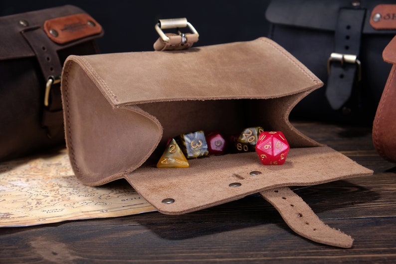 May include: A light brown leather pouch with a buckle closure containing several multi-sided gaming dice. The pouch is open, revealing a red twenty-sided die in the foreground and other assorted dice inside, including yellow and marbled patterns.