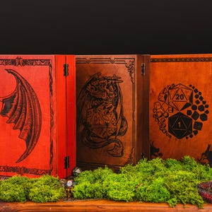 May include: Three wooden boxes with hinged lids, featuring engraved fantasy-themed designs. The boxes are in shades of red and brown, with images of a dragon wing, a dragon, and a D20 die. The boxes are displayed on a wooden surface with green moss.