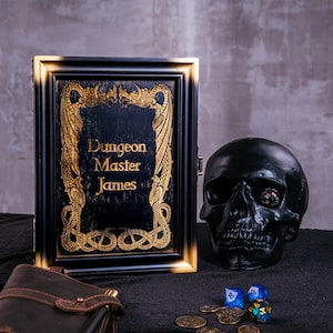 May include: A black and gold framed box with the text "Dungeon Master James" is displayed next to a black skull, dice, and coins. The box has ornate gold detailing. The skull has a die in its eye socket.