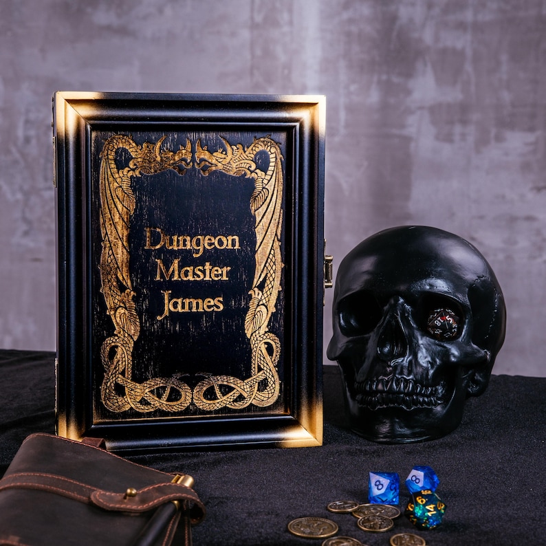 May include: A black and gold framed box with the text "Dungeon Master James" is displayed next to a black skull, dice, and coins. The box has ornate gold detailing. The skull has a die in its eye socket.