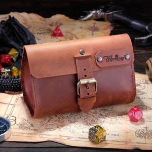 May include: A brown leather pouch with a front flap, a metal buckle closure, and a rectangular leather patch embossed with the text "CRIT HAPPENS" secured by two metal rivets. The pouch features visible stitching along the edges and a smooth finish.
