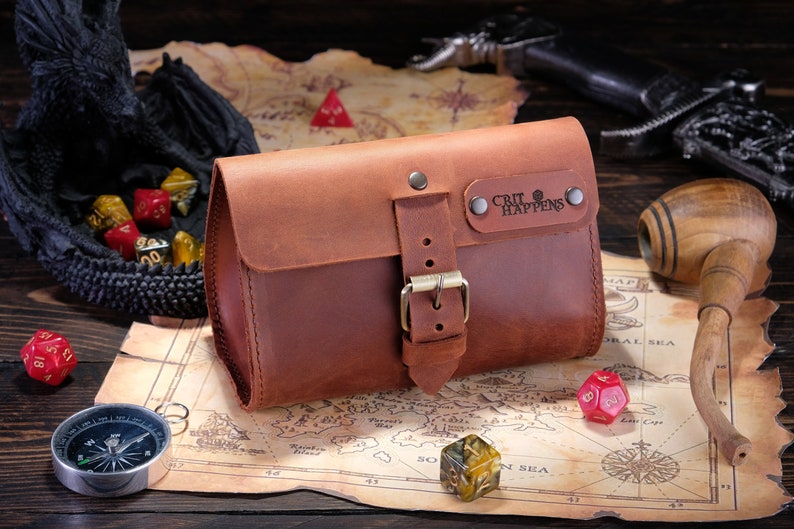 May include: A brown leather pouch with a front flap, a metal buckle closure, and a rectangular leather patch embossed with the text "CRIT HAPPENS" secured by two metal rivets. The pouch features visible stitching along the edges and a smooth finish.