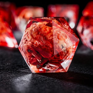 Puede incluir: Un dado de veinte caras con una cubierta de resina roja transl&uacute;cida que encierra un inserto texturizado de color beige y rojo similar a un cerebro. El dado tiene bordes afilados y n&uacute;meros grabados claros, con otros dados similares visibles al fondo.