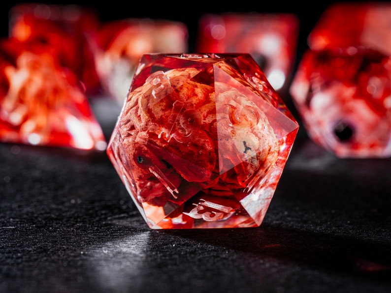 Puede incluir: Un dado de veinte caras con una cubierta de resina roja transl&uacute;cida que encierra un inserto texturizado de color beige y rojo similar a un cerebro. El dado tiene bordes afilados y n&uacute;meros grabados claros, con otros dados similares visibles al fondo.