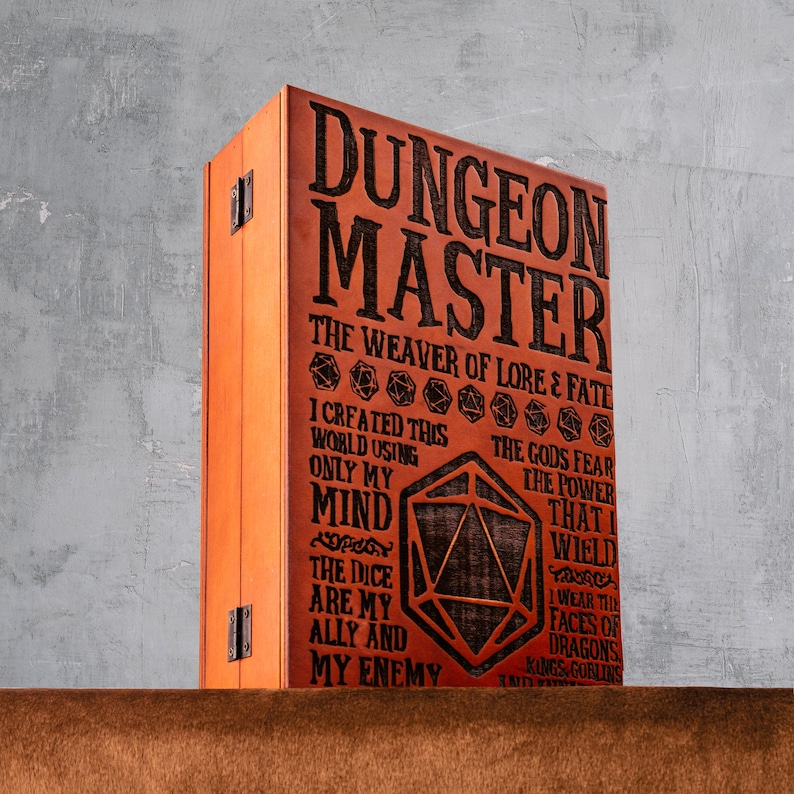 May include: A rectangular wooden box with a light brown finish and black metal hinges. The front panel features the engraved text DUNGEON MASTER THE WEAVER OF LORE & FATE, along with various dice icons and a large 20-sided die graphic in the center.