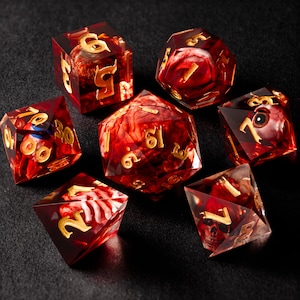 Op de afbeelding: Set van zeven rode en gouden dobbelstenen met verschillende symbolen en ontwerpen. De dobbelstenen zijn gemaakt van helder hars met een draaiend rood en gouden patroon. De dobbelstenen zijn genummerd van 1 tot en met 6.