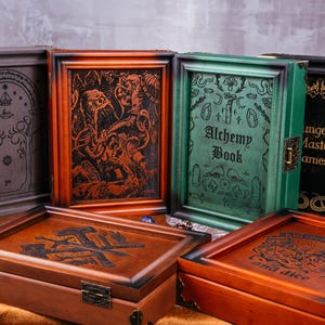 May include: Wooden game boxes with engraved designs. The boxes are various colors, including brown, green, and black. Some boxes have text that reads "Alchemy Book" and "Dungeon Master Games". The boxes are designed for board games.