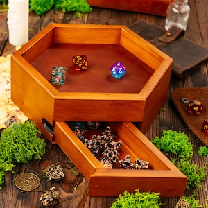 Puede incluir: Una bandeja hexagonal de madera para dados con una base de cuero marrón oscuro, que cuenta con un cajón extraíble lleno de varios dados de juego multifacéticos. Tres dados adicionales están colocados sobre la superficie de la bandeja, incluyendo opciones en rojo, azul y verde.