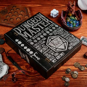 Peut inclure: Une bo&icirc;te en bois noire "Dungeon Master" avec des lettres blanches et des graphiques de d&eacute;s. La bo&icirc;te est entour&eacute;e d'objets li&eacute;s au jeu, notamment des pi&egrave;ces de monnaie, une figurine de dragon et un plateau de d&eacute;s en bois.