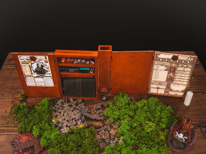 Peut inclure: Un ensemble de jeu de r&ocirc;le de table en bois avec une finition marron. L'ensemble comprend un compartiment de rangement pour d&eacute;s, un portefeuille en simili cuir et divers accessoires de jeu. Un paysage de mousse verte et des cartes de jeu sont au premier plan.
