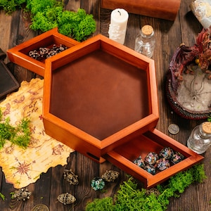 Puede incluir: Una bandeja hexagonal de madera para dados con una superficie de rodadura de cuero marrón liso y un cajón extraíble que contiene varios dados poliédricos. La bandeja está construida en madera pulida con un acabado marrón rojizo.