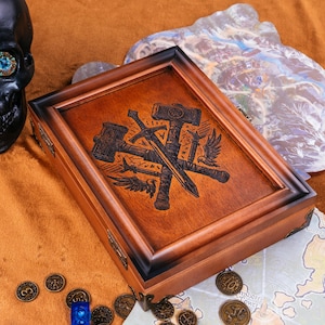 May include: A wooden box with a dark brown frame and a lighter brown interior. The box features an engraved design of a sword, hammers, and wings. The box is surrounded by coins and a map.