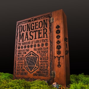 Peut inclure: Un coffret en bois marron avec l'inscription "DUNGEON MASTER" grav&eacute;e. Le coffret pr&eacute;sente des symboles de d&eacute;s et du texte, dont "THE WEAVER OF LORE & FATE". Il est pos&eacute; sur de la mousse verte.