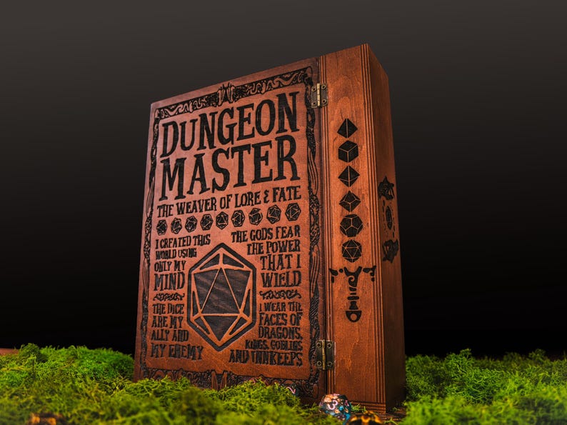 Peut inclure: Un coffret en bois marron avec l'inscription "DUNGEON MASTER" grav&eacute;e. Le coffret pr&eacute;sente des symboles de d&eacute;s et du texte, dont "THE WEAVER OF LORE & FATE". Il est pos&eacute; sur de la mousse verte.
