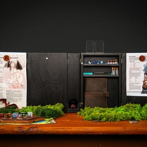 Puede incluir: Un organizador de juegos de mesa de madera negra con múltiples compartimentos, que incluyen un armario central, estantes y una torre de dados. El organizador contiene dados, un cuaderno y hojas de personajes impresas. Musgo verde y un tablero de juego de madera están sobre la mesa.