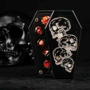 Puede incluir: Una caja negra con forma de ata&uacute;d que presenta un dise&ntilde;o grabado de cuatro calaveras apiladas en la tapa. La caja est&aacute; parcialmente abierta, revelando un juego de cinco dados poli&eacute;dricos rojos transl&uacute;cidos con n&uacute;meros blancos dentro.