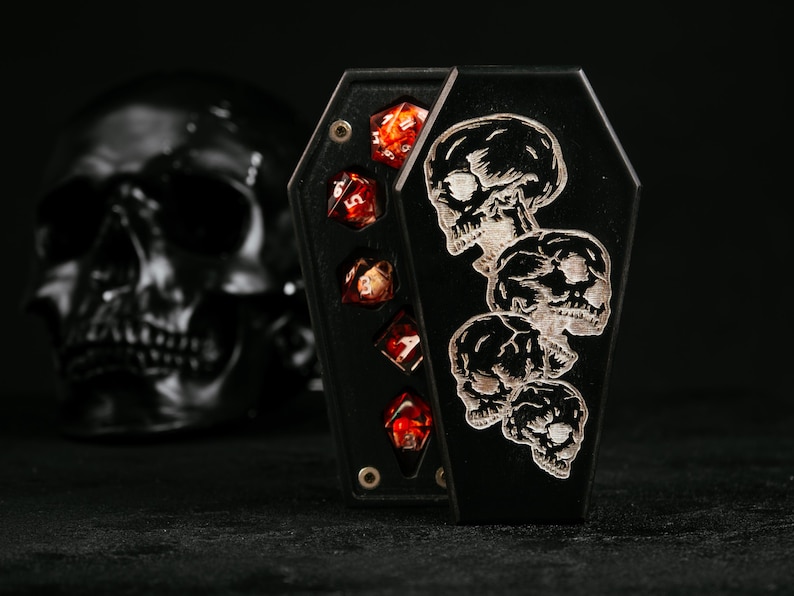 Puede incluir: Una caja negra con forma de ata&uacute;d que presenta un dise&ntilde;o grabado de cuatro calaveras apiladas en la tapa. La caja est&aacute; parcialmente abierta, revelando un juego de cinco dados poli&eacute;dricos rojos transl&uacute;cidos con n&uacute;meros blancos dentro.