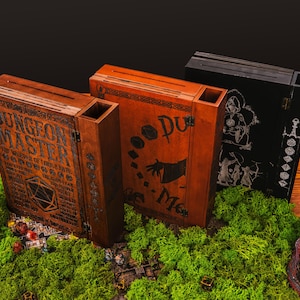 Peut inclure: Trois bo&icirc;tes en bois de Ma&icirc;tre du Donjon avec des motifs et du texte grav&eacute;s. Les bo&icirc;tes sont marron, orange et noires, et reposent sur un lit de mousse verte. Les bo&icirc;tes ont une fente sur le dessus et sont d&eacute;cor&eacute;es d'illustrations et de texte sur le th&egrave;me de la fantaisie.