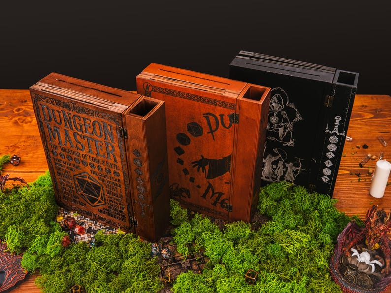 Peut inclure: Trois bo&icirc;tes en bois de Ma&icirc;tre du Donjon avec des motifs et du texte grav&eacute;s. Les bo&icirc;tes sont marron, orange et noires, et reposent sur un lit de mousse verte. Les bo&icirc;tes ont une fente sur le dessus et sont d&eacute;cor&eacute;es d'illustrations et de texte sur le th&egrave;me de la fantaisie.