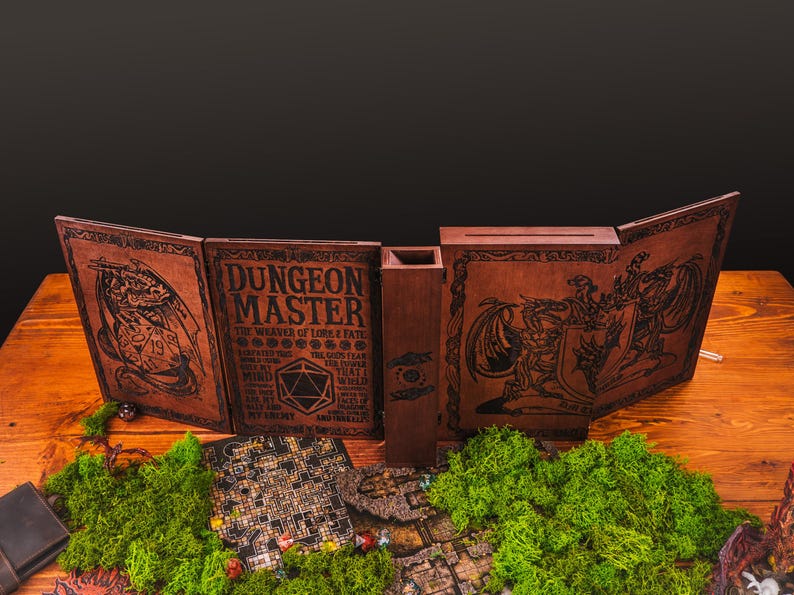 Peut inclure: Un &eacute;cran de ma&icirc;tre de donjon en bois marron avec des illustrations fantastiques grav&eacute;es et le texte "DUNGEON MASTER". L'&eacute;cran est ouvert, r&eacute;v&eacute;lant un bac &agrave; d&eacute;s et une carte de jeu miniature. De la mousse verte et des pi&egrave;ces de jeu sont au premier plan.
