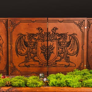 May include: A wooden game master screen with a dragon crest design. The screen is brown with black engraved artwork. The center panel features a dragon crest with the text "DM Thomas". The screen is decorated with green moss and dice.